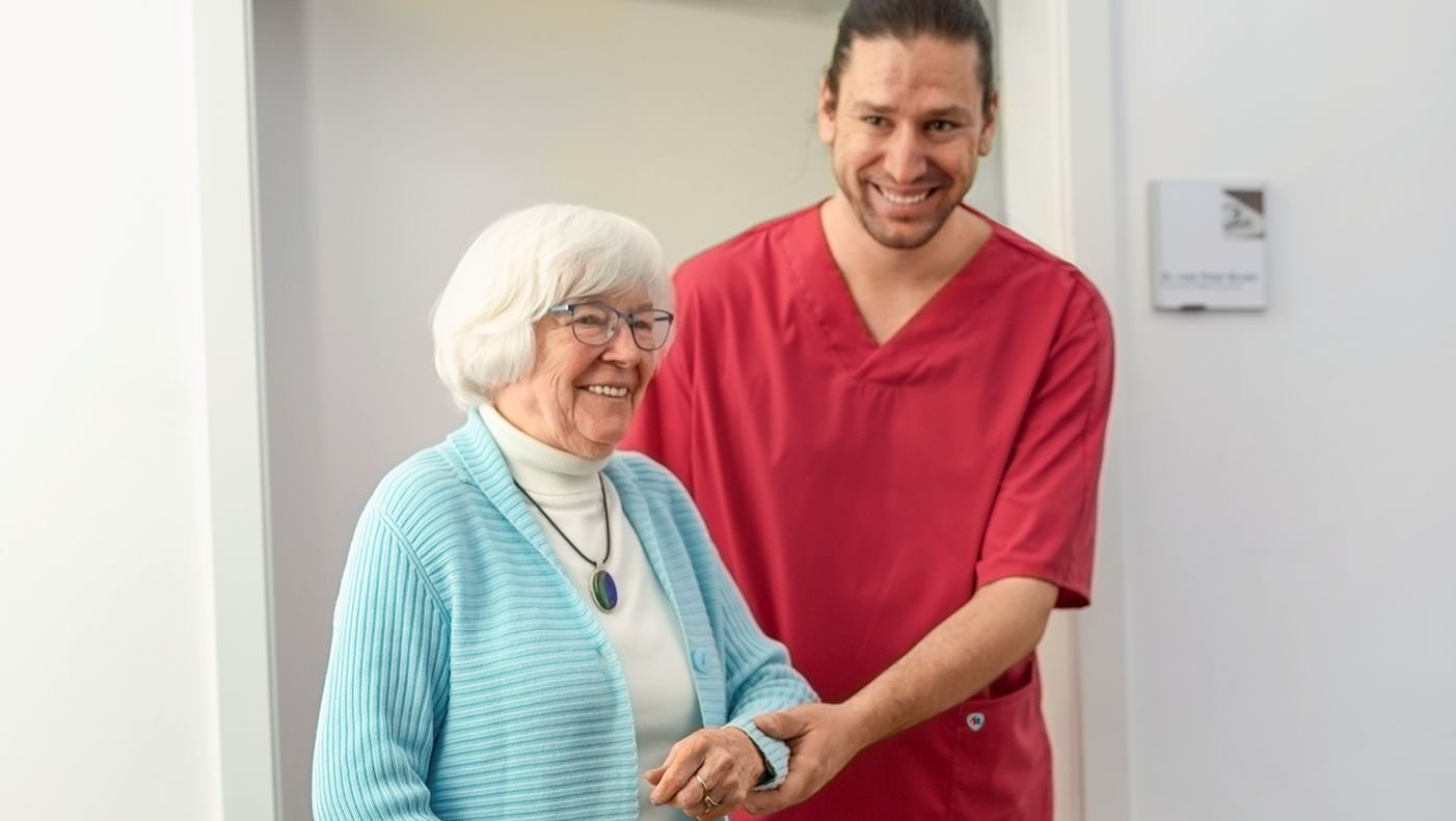 Elderly care worker in a red coat helps a senior citizen to walk.