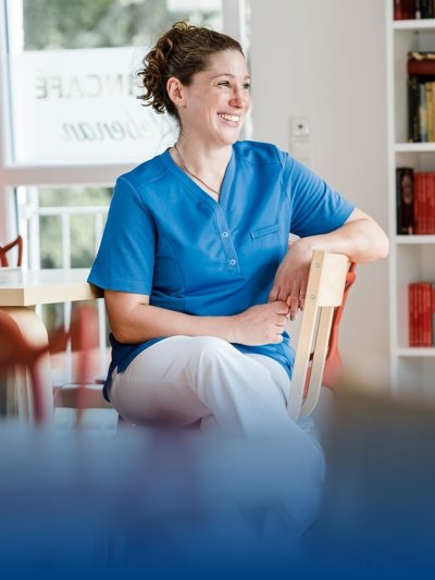 A carer wears a blue tunic and sits in a chair with a smile.