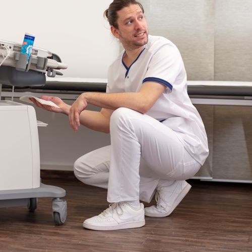 A nurse is in a kneeling position and wears white work trousers and a tunic.