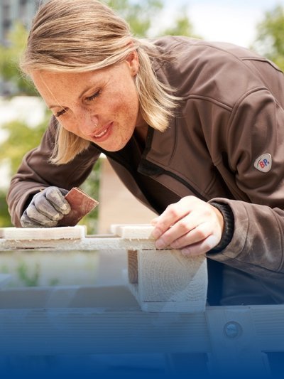 A carpenter wears brown work clothes and sands wood.